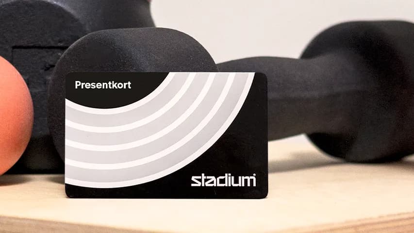 A gym equipment setup with a black dumbbell, orange ball, and pink yoga mat on a wooden surface. A gift card reads "Presentkort" and "stadium."